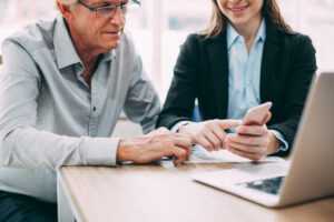 Smiling young businesswoman showing smartphone screen to colleague. They sitting at table and discussing business project. They using phone together to find necessary information. Teamwork concept